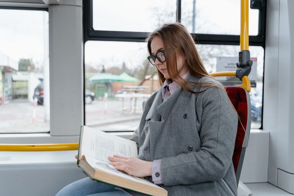 A woman in a gray coat is reading a book while on a bus.