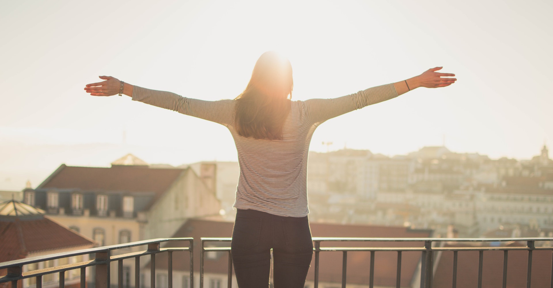 A woman with outstretched arms looking out towards the sunset while standing on a balcony.